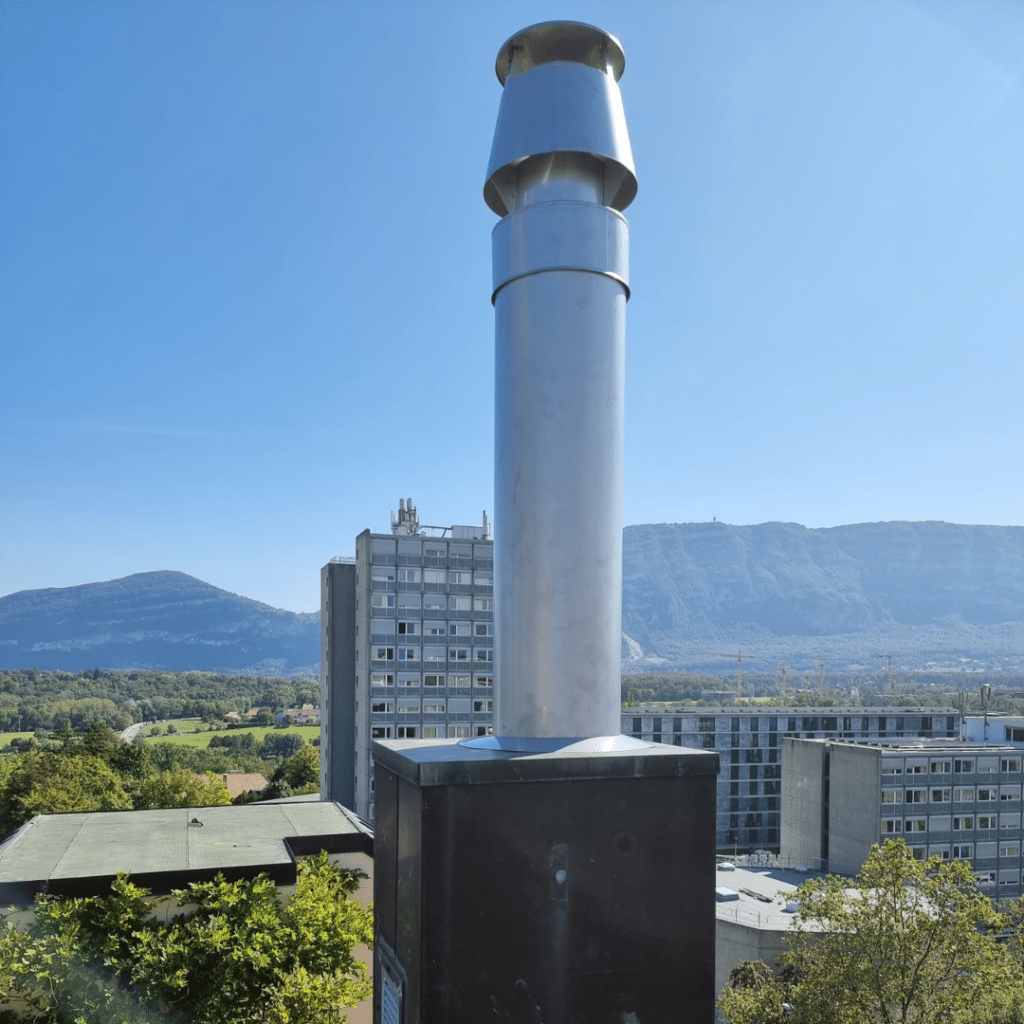 Conduit de cheminée en acier inoxydable avec vue sur des bâtiments et des montagnes en arrière-plan.