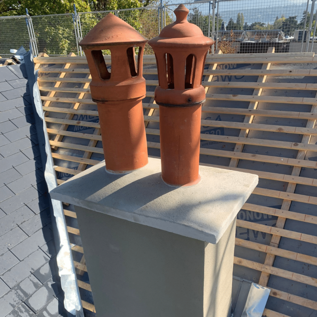 Deux chapeaux de cheminée en terre cuite installés sur un conduit en béton pendant une rénovation de toiture.